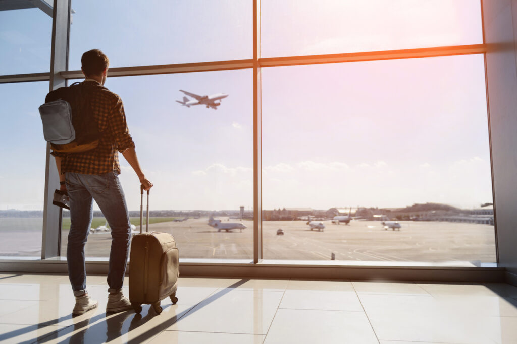 Ein Mann mit Koffer und Rucksack sieht seinen verpassten Anschlussflug starten, Rückenansicht.