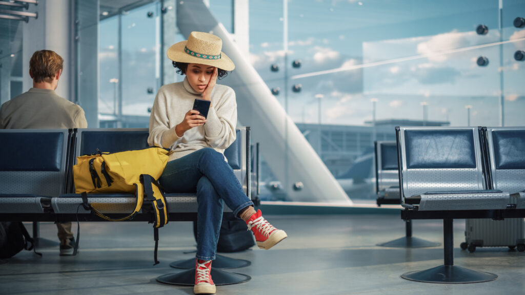 Frau mit Urlaubshut sitzt am Flughafengate und erhält die schlechte Nachricht, dass ihr Flug ausgefallen ist.